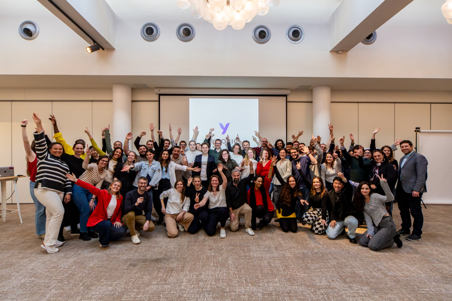 YOOBIC teammates posing together in an auditorium at the March 2025 boot camp, some kneeling in front and others cheering with raised hands